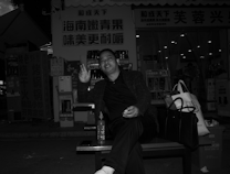 A person sitting on a bench outside a store with bright signage. The individual is smiling and gesturing with their hand, while a bottle and two bags are placed beside them. The background includes illuminated items inside a shop, with price tags visible.