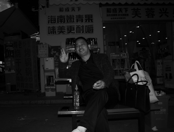 A person sitting on a bench outside a store with bright signage. The individual is smiling and gesturing with their hand, while a bottle and two bags are placed beside them. The background includes illuminated items inside a shop, with price tags visible.