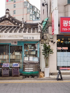 A traditional Korean building with a tiled roof housing a small bookstore. The storefront is filled with books and some are displayed openly. Nearby, a modern building contrasts with the rustic structure. A potted plant stands in front of the shop, and signage with Korean writing is visible, suggesting this is a street scene in an urban area.