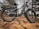 A black fat-tire electric bike is positioned on a rocky terrain surrounded by pine trees. The bike features large, rugged tires, a sturdy frame, and a visible chain mechanism. The brand name 'DENAGO' is printed on the bike's body.
