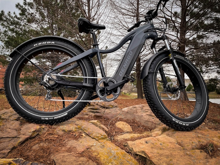 A black fat-tire electric bike is positioned on a rocky terrain surrounded by pine trees. The bike features large, rugged tires, a sturdy frame, and a visible chain mechanism. The brand name 'DENAGO' is printed on the bike's body.