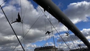Silhouettes of acrobatic sculptures are suspended on a bridge, performing balancing acts on ropes and metal structures against a backdrop of a partly cloudy blue sky.