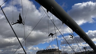Silhouettes of acrobatic sculptures are suspended on a bridge, performing balancing acts on ropes and metal structures against a backdrop of a partly cloudy blue sky.