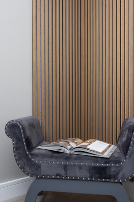 A plush, gray chaise lounge with metallic studs along its edges is positioned against a wall adorned with vertical wooden slats. An open magazine rests on the seat, displaying colorful pages.