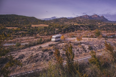 A motorhome is parked on a rugged, hilly landscape surrounded by scattered trees and shrubbery. In the background, there are lush green hills and distant mountains under a partly cloudy sky.