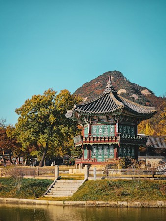 A traditional Korean pavilion set amidst autumn foliage, with a serene pond in the foreground and a mountain in the background. The structure features intricate architectural details with colorful wooden patterns, showcasing the historical design characteristic of Korean culture.