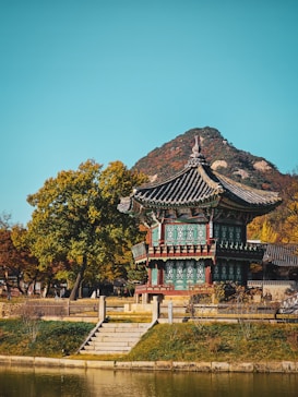 A traditional Korean pavilion set amidst autumn foliage, with a serene pond in the foreground and a mountain in the background. The structure features intricate architectural details with colorful wooden patterns, showcasing the historical design characteristic of Korean culture.