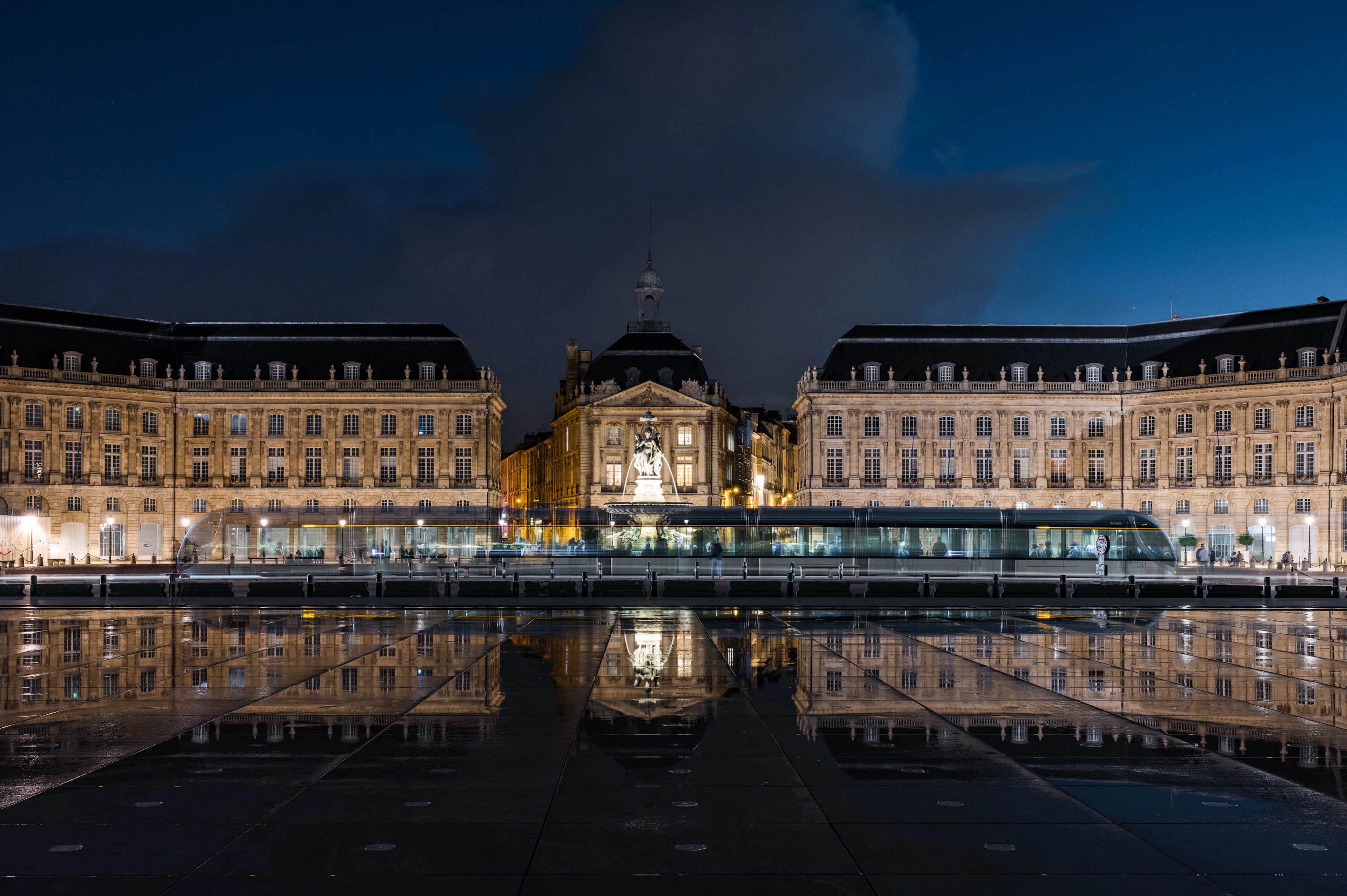The floodlit 18th-century facades of the Place de la Bourse in Bordeaux mirrored in the wet stone of the Miroir d'Eau at blue hour, a lit tram crossing the quay in front
