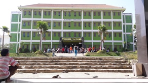 A large, green university building with multiple stories, featuring the sign 'Fakultas Tarbiyah' above the entrance. People are gathered on the steps in front of the building, some sitting while others stand. The scene is framed by small palm trees and stone steps leading up to the entrance.