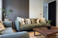 A modern living room featuring a green velvet sofa adorned with numerous decorative cushions, alongside a matching armchair. A wooden coffee table is placed in front, and a small potted plant adds a touch of nature. The walls are decorated with a striped textured panel, creating a stylish backdrop. A black door is slightly open, leading to another room.