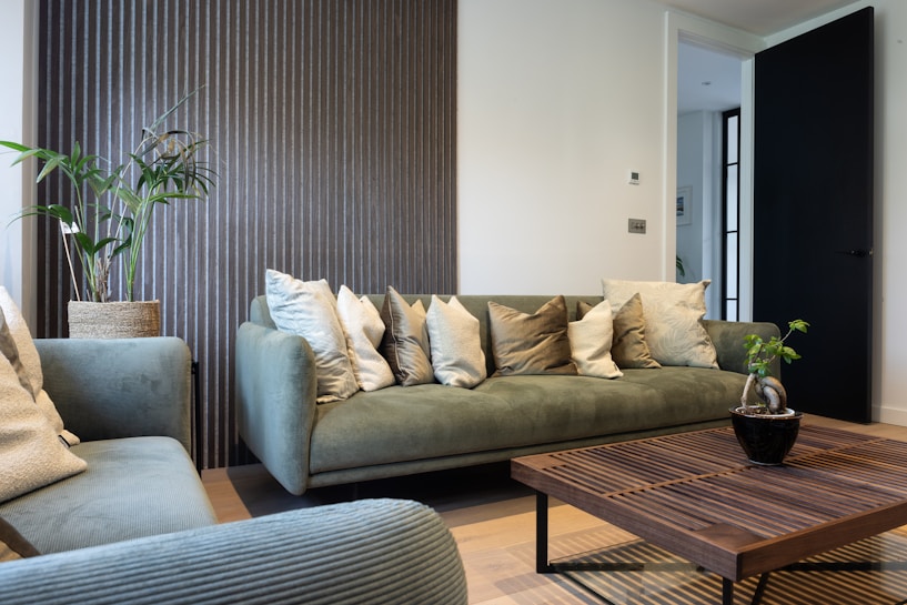 A modern living room featuring a green velvet sofa adorned with numerous decorative cushions, alongside a matching armchair. A wooden coffee table is placed in front, and a small potted plant adds a touch of nature. The walls are decorated with a striped textured panel, creating a stylish backdrop. A black door is slightly open, leading to another room.