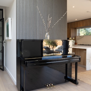 A black piano stands elegantly against a textured vertical striped wall. A vase with long branches adds a touch of nature above the piano. The adjacent room, visible through an open doorway, features wooden cabinets, a light-filled window, and a modern chair with sunlight streaming in.