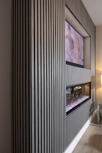 A modern interior featuring a wall with vertical wooden slats, a recessed television, and a contemporary fireplace. The TV displays an unclear image of two people, while the fireplace below emits a glowing light from artificial logs. The setting exudes a sleek and stylish design, with subtle lighting enhancing the atmosphere. A small glass side table with a lamp adds to the minimalist decor.