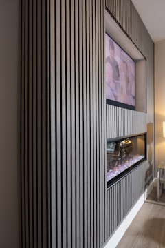 A modern interior featuring a wall with vertical wooden slats, a recessed television, and a contemporary fireplace. The TV displays an unclear image of two people, while the fireplace below emits a glowing light from artificial logs. The setting exudes a sleek and stylish design, with subtle lighting enhancing the atmosphere. A small glass side table with a lamp adds to the minimalist decor.