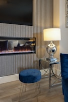 A cozy room interior featuring a modern electric fireplace with a flat-screen TV above it. The setup is complemented by a sleek glass lamp with a white shade on a glass side table, casting a warm glow. A blue velvet stool and chair add a touch of elegance, alongside a light wood floor, and vertical textured wall panels.