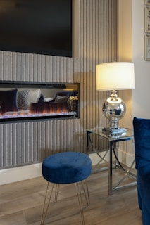 A cozy room interior featuring a modern electric fireplace with a flat-screen TV above it. The setup is complemented by a sleek glass lamp with a white shade on a glass side table, casting a warm glow. A blue velvet stool and chair add a touch of elegance, alongside a light wood floor, and vertical textured wall panels.