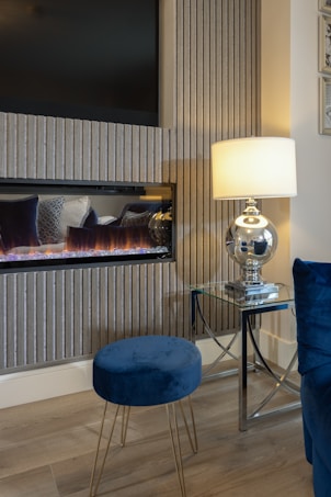 A cozy room interior featuring a modern electric fireplace with a flat-screen TV above it. The setup is complemented by a sleek glass lamp with a white shade on a glass side table, casting a warm glow. A blue velvet stool and chair add a touch of elegance, alongside a light wood floor, and vertical textured wall panels.