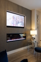 A modern living room setup with vertical wooden paneling, featuring a wall-mounted television and a sleek, horizontal electric fireplace beneath it. To the right, there is a glass side table with a metallic lamp that has a reflective, silver finish. The room has a cozy atmosphere, enhanced by the warm glow of the fireplace. Several framed pictures are displayed on the wall, adding a decorative touch.