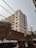 A tall, rectangular residential building with multiple balconies and numerous windows. The structure is surrounded by power lines crisscrossing the scene, and there's a brick wall topped with barbed wire in the foreground. The sky is clear, and a man is visible in the lower right corner, standing near a white vehicle and an auto-rickshaw.