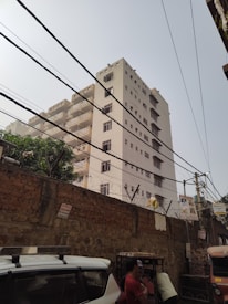 A tall, rectangular residential building with multiple balconies and numerous windows. The structure is surrounded by power lines crisscrossing the scene, and there's a brick wall topped with barbed wire in the foreground. The sky is clear, and a man is visible in the lower right corner, standing near a white vehicle and an auto-rickshaw.