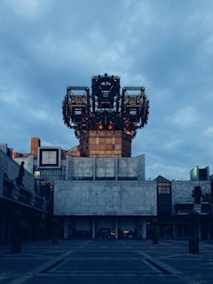 A large, industrial-style building with a futuristic, geometrical top structure. The facade is mostly concrete with rectangular and square patterns, while the sky above is overcast with gray clouds.