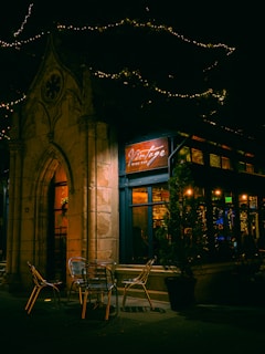 An outdoor seating area of a quaint wine bar is depicted. Metal chairs and a table are placed on the pavement, under warm ambient light. The entrance has a stone archway, and string lights are hanging above. A sign reading 'Vintage Wine Bar' is illuminated, adding a cozy glow to the scene.