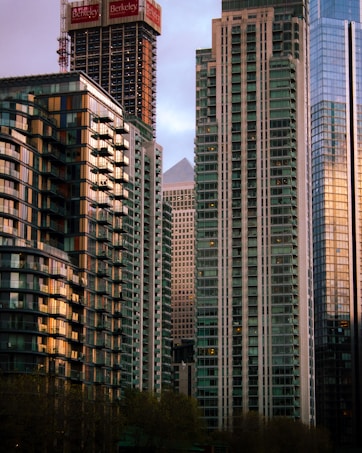 A cluster of modern skyscrapers with a mix of architecture styles, featuring reflective glass and geometric designs. The buildings rise closely together against a pastel sky, with some construction visible on the rooftops.