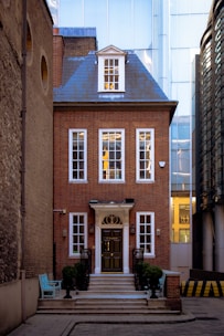 A charming red brick townhouse set in a narrow alleyway, featuring tall white-framed windows and a central dark door accessed by a staircase with black railings. The building has a decorative gable roof with a window, and it contrasts with the modern glass-faced buildings surrounding it. A turquoise bench is placed to the side, adding a pop of color to the scene.