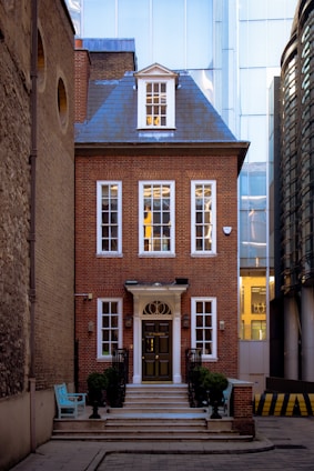A charming red brick townhouse set in a narrow alleyway, featuring tall white-framed windows and a central dark door accessed by a staircase with black railings. The building has a decorative gable roof with a window, and it contrasts with the modern glass-faced buildings surrounding it. A turquoise bench is placed to the side, adding a pop of color to the scene.