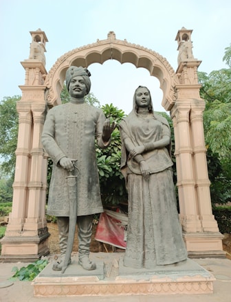 A pair of statues representing a man and a woman dressed in traditional attire stand beneath a decorative arch. The man is wearing a turban and holding a sword, while the woman is adorned in a sari with jewelry. The arch is ornately carved, and there is lush greenery in the background.