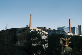 An industrial building with tall brick smokestacks standing prominently against a clear blue sky. In the foreground, there is a mix of trees and greenery partially obscuring the structure. Modern office buildings are visible in the background.