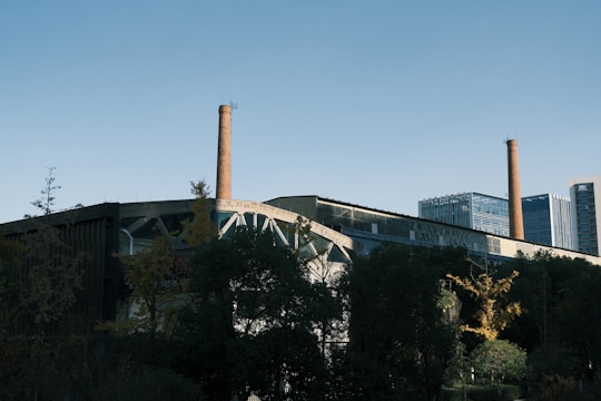 An industrial building with tall brick smokestacks standing prominently against a clear blue sky. In the foreground, there is a mix of trees and greenery partially obscuring the structure. Modern office buildings are visible in the background.
