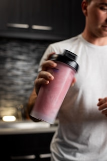 A person in a white shirt is holding and shaking a pink smoothie or protein shake in a large cup with a black lid. The background shows a kitchen setting with dark cabinets and a tiled backsplash.
