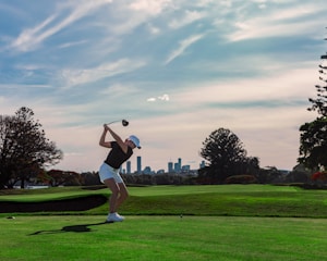 A person is in the midst of a golf swing on a sprawling golf course, with a city skyline visible in the distance under a partly cloudy sky. The lush green grass contrasts with the silhouette of the player, who is wearing a white cap and shorts.