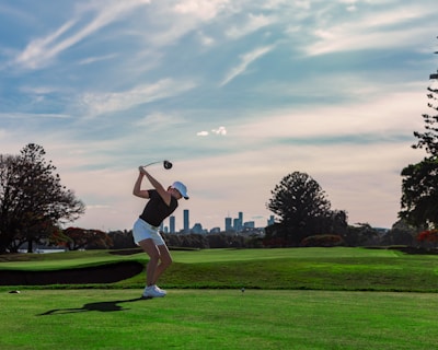 A person is in the midst of a golf swing on a sprawling golf course, with a city skyline visible in the distance under a partly cloudy sky. The lush green grass contrasts with the silhouette of the player, who is wearing a white cap and shorts.
