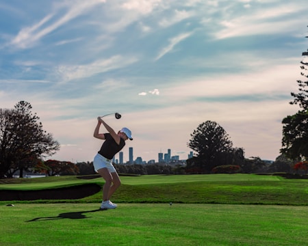 A person is in the midst of a golf swing on a sprawling golf course, with a city skyline visible in the distance under a partly cloudy sky. The lush green grass contrasts with the silhouette of the player, who is wearing a white cap and shorts.