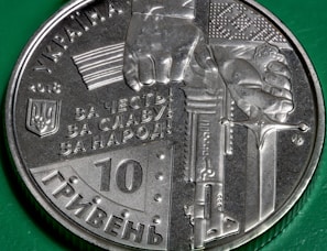 A Ukrainian coin from 2018 with a design that includes a hand gripping a sword and a block of text in Cyrillic. The coin has intricate detailing, including the Ukrainian coat of arms and a denomination of 10 hryvnias. The background features a green surface.