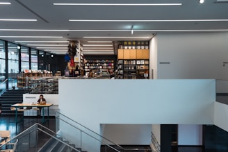 A large, modern indoor space featuring a multi-level bookstore or library. Books and magazines are neatly arranged on shelves, and a person is sitting at a table working on a laptop. The design is sleek, with clean lines and contemporary lighting.