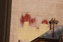 A textured wall features faded red paint marks that resemble flames. Below the wall is a metal roof with visible rusty spots and two chimney stacks, one of which is connected to a red pipe. The color contrast between the wall and roof creates an industrial, urban atmosphere.