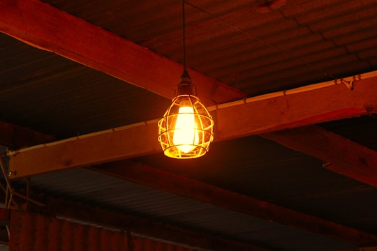 A single incandescent light bulb enclosed within a metal cage hangs from a wooden beam ceiling. The warm yellow glow of the bulb contrasts with the darker, rustic look of the wooden beams and the corrugated metal ceiling. Electrical wiring is visible running along the beams.