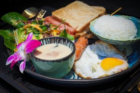 A well-presented meal consists of a variety of food items arranged on a plate. There is a bowl of rice, a cup of creamy soup with a slice of mushroom, a fried egg with a runny yolk, slices of toast, a mixed green salad with a slice of smoked salmon, and a piece of grilled meat on a skewer. A purple orchid flower is placed on the salad for decoration.