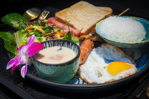 A well-presented meal consists of a variety of food items arranged on a plate. There is a bowl of rice, a cup of creamy soup with a slice of mushroom, a fried egg with a runny yolk, slices of toast, a mixed green salad with a slice of smoked salmon, and a piece of grilled meat on a skewer. A purple orchid flower is placed on the salad for decoration.