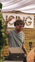 A DJ is performing at an outdoor event, wearing headphones and a gray t-shirt, with one hand raised. Behind him is a bamboo backdrop with a partially visible white sign. Plants are hanging on the left side of the image.