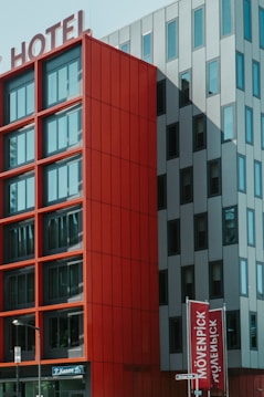 A modern hotel building with a striking facade featuring large red panels and a contrasting gray section with vertical window arrangements. The signage at the top indicates it is a hotel, while street signs are visible at ground level.