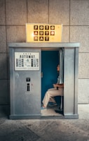 A vintage photo booth with the word 'FOTOAUTOMAT' displayed prominently at the top. The booth is metallic and looks slightly worn. Inside, a person is seated, partially visible with a blue interior background. The floor is stone or concrete, and there are some faint graffiti marks on the booth's exterior.