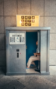 A vintage photo booth with the word 'FOTOAUTOMAT' displayed prominently at the top. The booth is metallic and looks slightly worn. Inside, a person is seated, partially visible with a blue interior background. The floor is stone or concrete, and there are some faint graffiti marks on the booth's exterior.