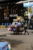 A street vendor is pushing a cart loaded with various household goods. The cart is stocked with colorful plastic baskets, brooms, and other miscellaneous items. The background features a store with a sign reading 'THE OASIS' and vibrant yellow and blue chairs are lined up outside.