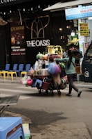 A street vendor is pushing a cart loaded with various household goods. The cart is stocked with colorful plastic baskets, brooms, and other miscellaneous items. The background features a store with a sign reading 'THE OASIS' and vibrant yellow and blue chairs are lined up outside.