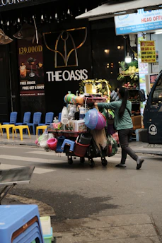 A street vendor is pushing a cart loaded with various household goods. The cart is stocked with colorful plastic baskets, brooms, and other miscellaneous items. The background features a store with a sign reading 'THE OASIS' and vibrant yellow and blue chairs are lined up outside.