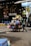 A street vendor is pushing a cart loaded with various household goods. The cart is stocked with colorful plastic baskets, brooms, and other miscellaneous items. The background features a store with a sign reading 'THE OASIS' and vibrant yellow and blue chairs are lined up outside.